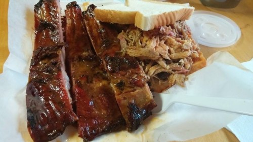 A plate of barbecue ribs and pulled pork served with white bread and a side of sauce.
