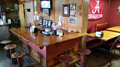A rustic BBQ restaurant interior featuring a wooden bar, stools, and Alabama memorabilia on the walls.