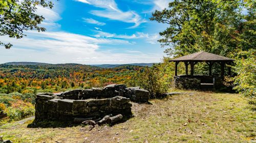 Scenic view of a landscape with colorful autumn trees, featuring a stone fire pit and a gazebo under a blue sky.