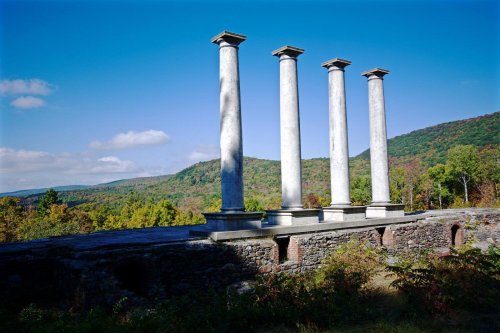 Four tall, weathered columns stand on a stone foundation, surrounded by colorful trees and a blue sky.