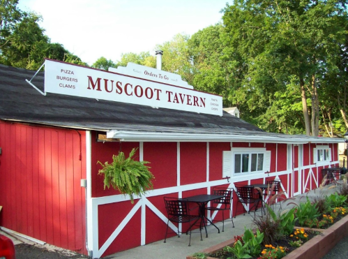 Exterior of Muscoot Tavern, a red building with a sign, outdoor seating, and a garden area.