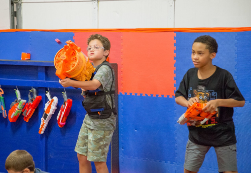 Two boys in a colorful play area hold toy blasters, ready for a fun game. Bright walls and equipment are in the background.