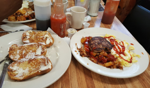 A plate of French toast with syrup and a hearty breakfast of eggs, potatoes, sausage, and ketchup on a wooden table.