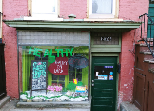 A storefront with a green sign reading "Healthy" and "Open," featuring plants and a chalkboard menu.