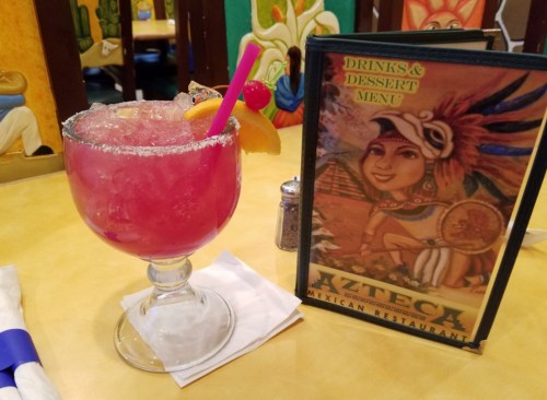 A colorful cocktail with a straw and fruit garnish next to a menu for Azteca Mexican Restaurant on a table.
