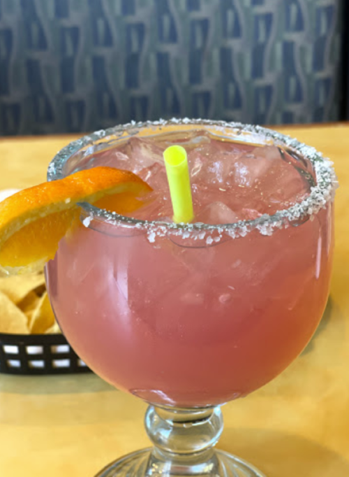 A colorful cocktail in a salt-rimmed glass, garnished with an orange slice and a green straw, next to a basket of chips.