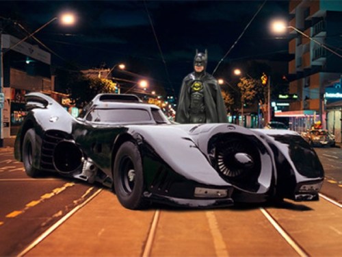 A superhero in a cape stands beside a sleek, futuristic black car on a city street at night.