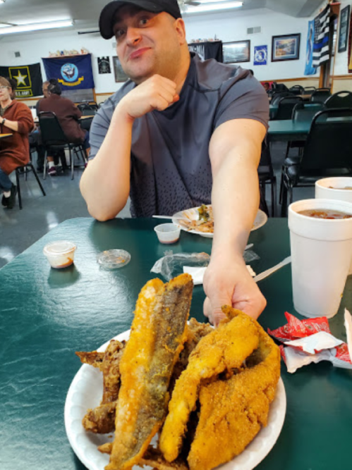 A man sitting at a table holds a plate of fried fish, with drinks and food in the background of a casual dining setting.