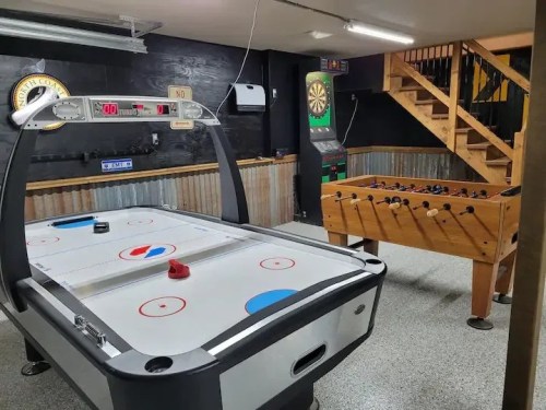 A game room featuring an air hockey table, foosball table, and a dartboard area with stairs in the background.