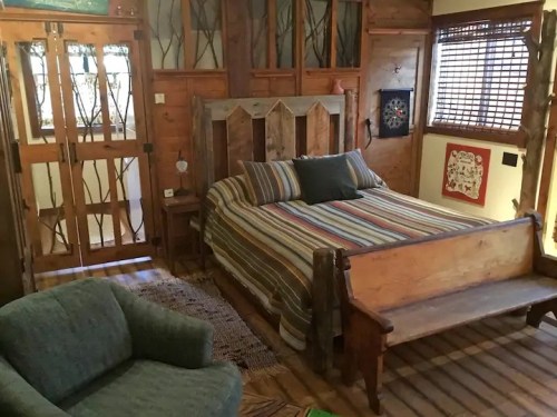 Cozy bedroom with a wooden bed, striped bedding, and rustic decor, featuring a green armchair and wooden accents.