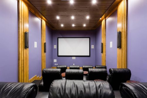 A cozy home theater with leather seating, purple walls, and a large screen at the front. Soft lighting enhances the ambiance.