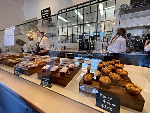 A bakery display with various pastries on wooden stands and staff behind a glass counter. Prices are visible.