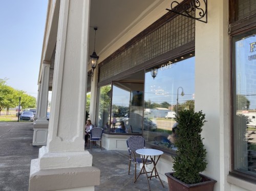 A café exterior with outdoor seating, large windows, and decorative plants, set on a sunny day.