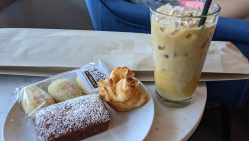 A plate with a chocolate cake, two pastries, and a tall glass of iced coffee on a marble table.