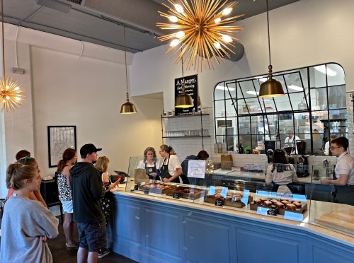 A busy café with customers in line, a display of pastries, and staff serving behind the counter. Modern decor and lighting.