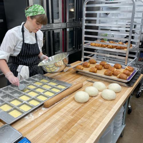 A baker in a kitchen prepares dough in trays, with rising dough and baked goods on a rack nearby.