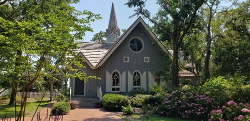 A charming gray house with a spire, surrounded by lush greenery and colorful flowers on a sunny day.