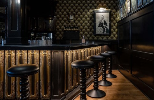 A stylish bar interior featuring black stools, ornate gold detailing, and vintage wallpaper with a framed portrait on the wall.