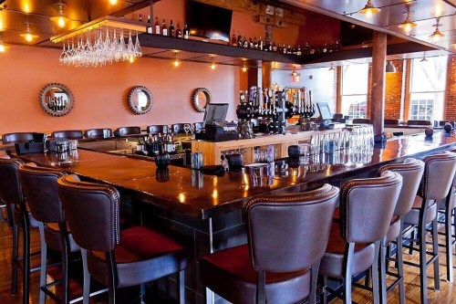 A stylish bar with a polished wooden counter, high stools, and warm lighting, featuring shelves of bottles and taps.