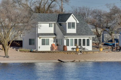 A cozy white house by the water, surrounded by trees and sandy beach, with a patio and chairs for relaxation.