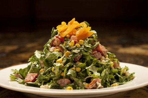 A vibrant salad with greens, corn, diced tomatoes, and orange slices, served on a white plate.