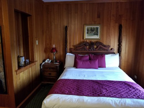 Cozy bedroom with wooden walls, a double bed with purple accents, a nightstand, and a small window.