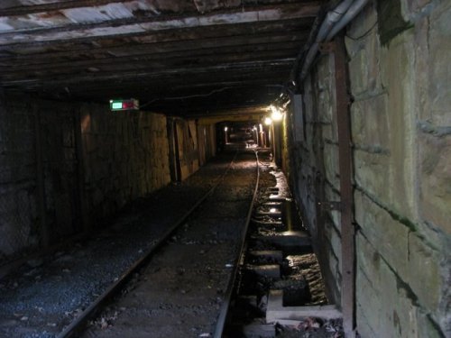 Dimly lit underground tunnel with stone walls, railway tracks, and puddles on the ground.