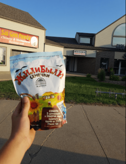 A hand holds a bag of sunflower seeds in front of a street with shops and a clear sky.