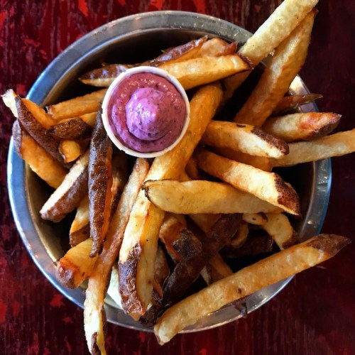 A bowl of golden-brown French fries with a small cup of purple dipping sauce in the center.