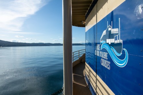 A view of a boat's side with a logo, reflecting on calm water under a clear blue sky.