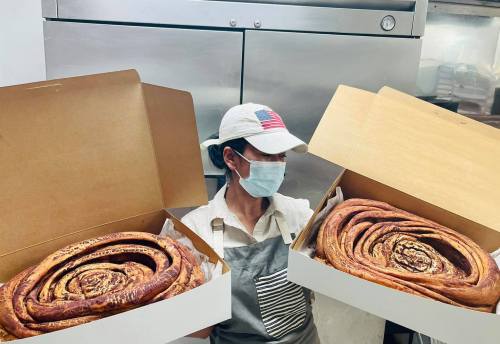 A person wearing a mask and cap holds two large boxes containing spiral pastries in a kitchen setting.