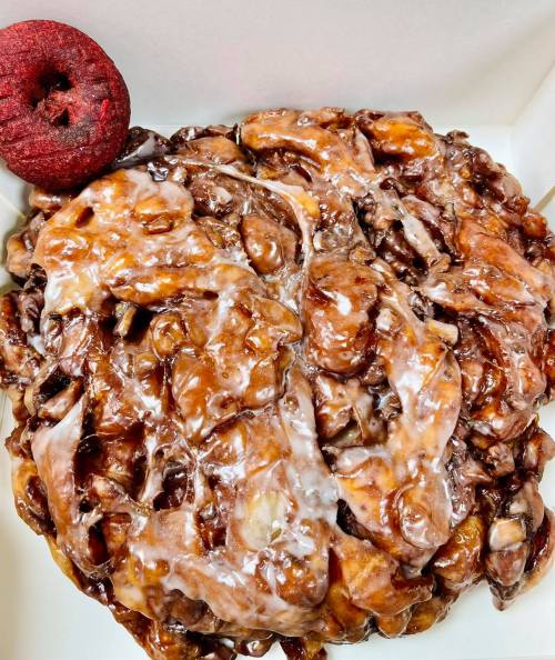 A close-up of a glazed apple fritter with a small red donut on the side, presented in a white box.