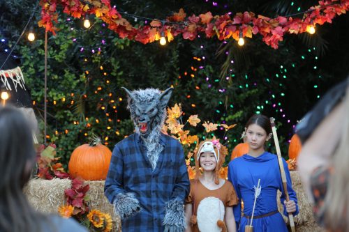 A werewolf-costumed person stands with two children in costumes, surrounded by autumn decorations and pumpkins.