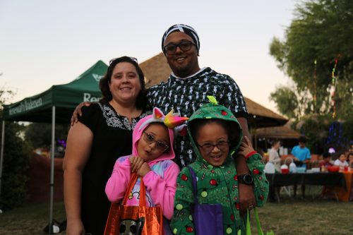 A family poses together outdoors, smiling, with two children in colorful costumes and adults in casual attire.