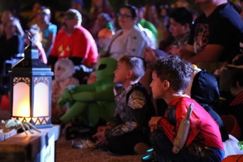 A group of children and adults in costumes watch a performance, with a lantern in the foreground and an alien figure behind.