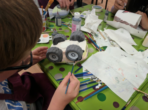 A child paints a toy car with gray wheels at a colorful table covered in art supplies and paper.