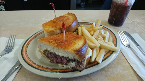 A grilled sandwich with meat and cheese, served with fries and a drink, on a plate with utensils beside it.