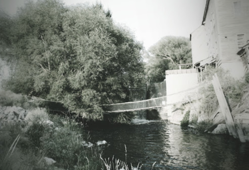 A black-and-white image of a river with a bridge, surrounded by trees and an old wooden structure.