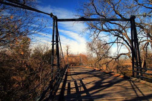 A wooden suspension bridge surrounded by bare trees under a clear blue sky. Shadows stretch across the bridge.
