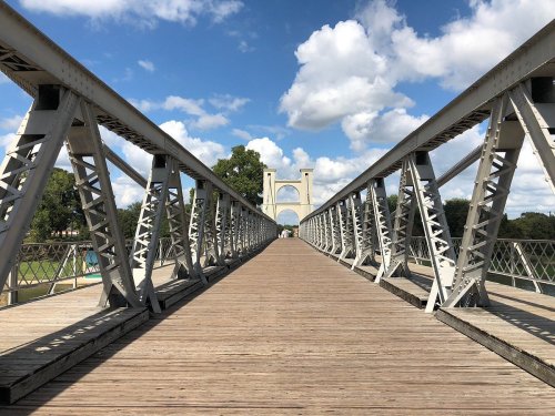 A wooden bridge with metal supports leads to an archway, under a blue sky with fluffy clouds.