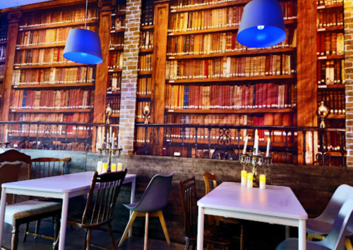 Cozy restaurant interior with white tables, colorful chairs, and a mural of bookshelves on the wall.