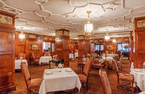Elegant restaurant interior with wooden paneling, ornate ceiling, and tables set with white linens and fine dining decor.