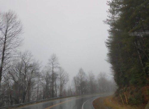 A winding road through a foggy landscape with bare trees and mist obscuring visibility.