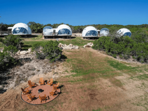 Aerial view of geodesic domes on a grassy area, with a circular seating arrangement around a fire pit in the foreground.