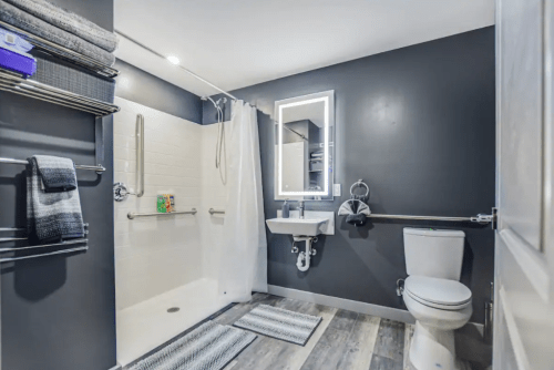 Modern bathroom featuring a shower, toilet, sink, and gray walls with towels and rugs.