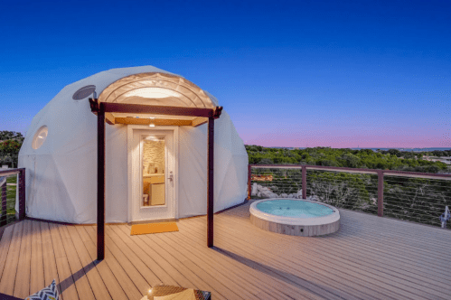 A modern dome structure with a hot tub on a wooden deck, set against a twilight sky.
