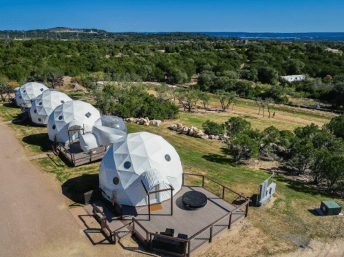 Aerial view of white geodesic domes on a wooden deck, surrounded by greenery and a clear blue sky.