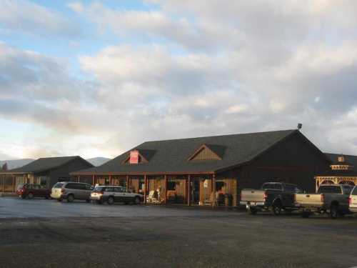 A rustic building with a dark exterior and a sloped roof, surrounded by parked vehicles under a cloudy sky.