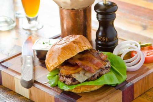 A juicy burger with bacon, cheese, lettuce, and tomato on a wooden board, accompanied by a knife and a drink in the background.