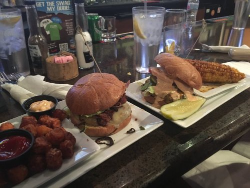 Two burgers with toppings, a side of tater tots with dipping sauce, and grilled corn on a bar counter.
